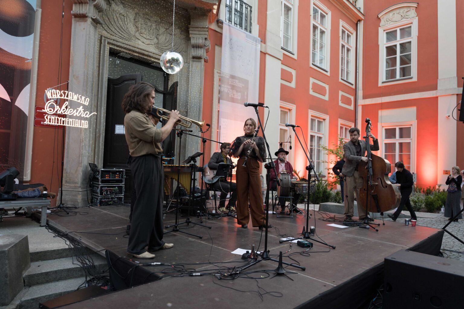 zespół na scenie, w tle czerwony budynek, nad wejściem do niego wisi dyskotekowa kula