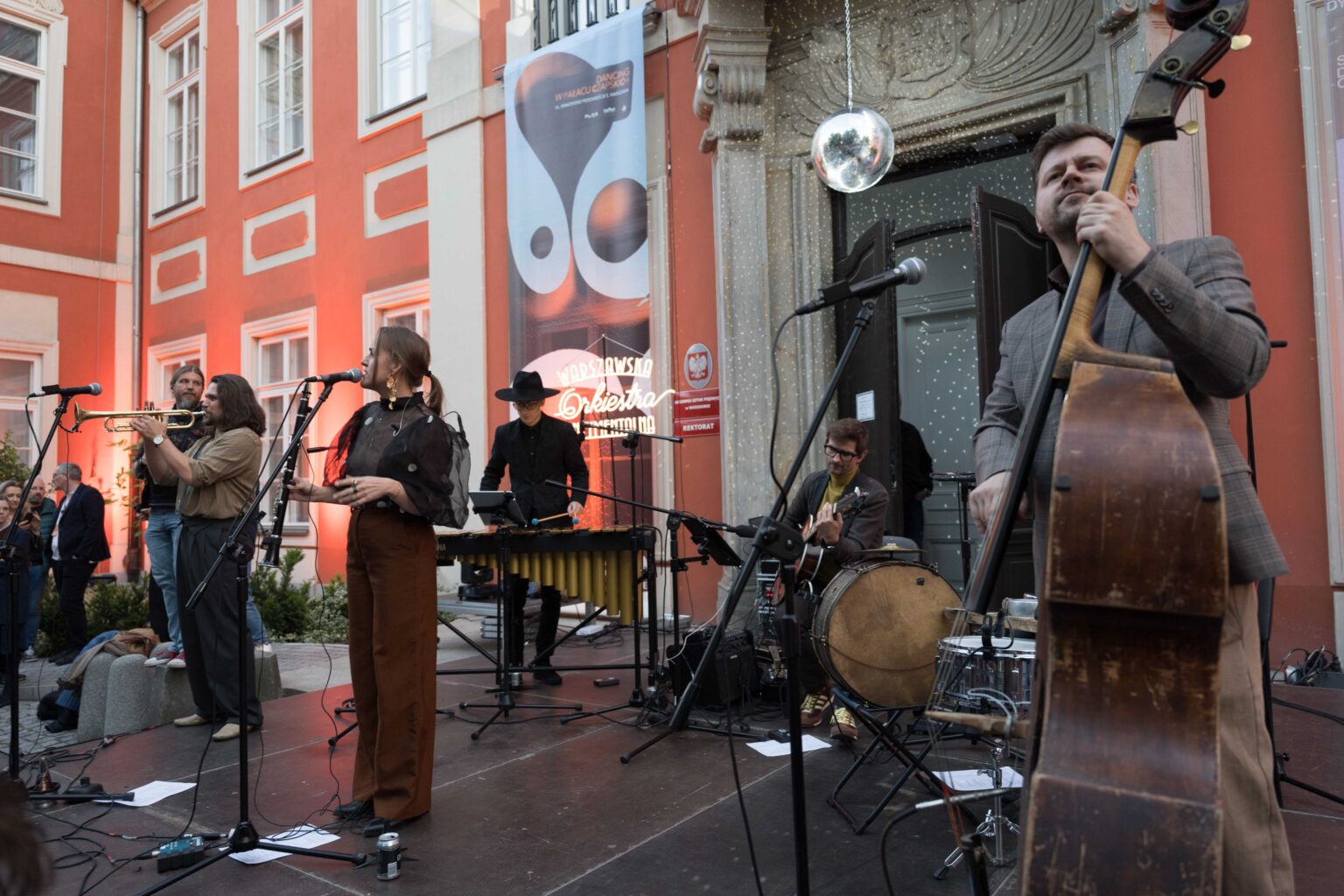 zespół na scenie, w tle czerwony budynek, nad wejściem do niego wisi dyskotekowa kula