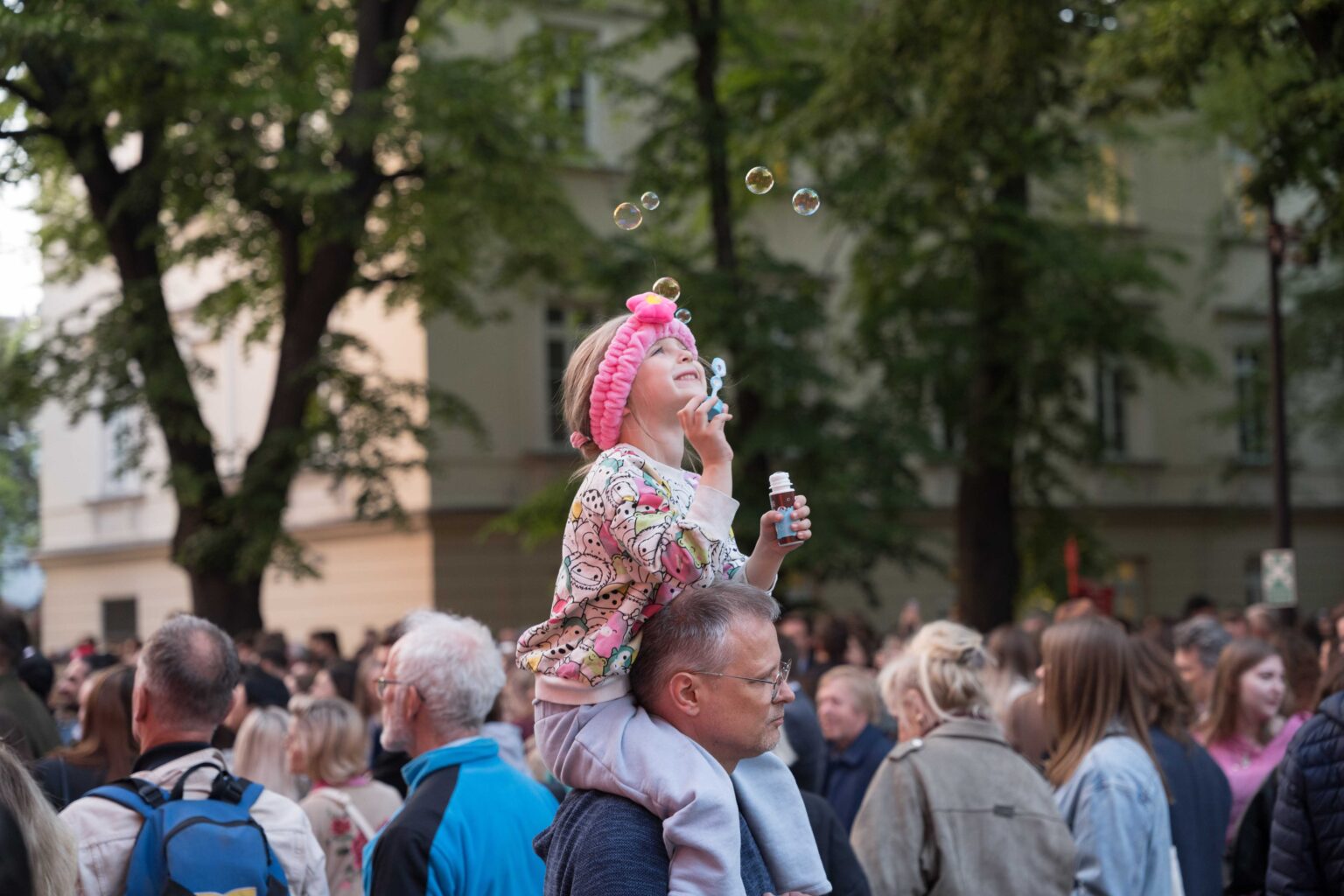 tłum ludzi na pierwszym planie mężczyz dziewczynką "na barana", dziewczynka puszcza bańki mydlane
