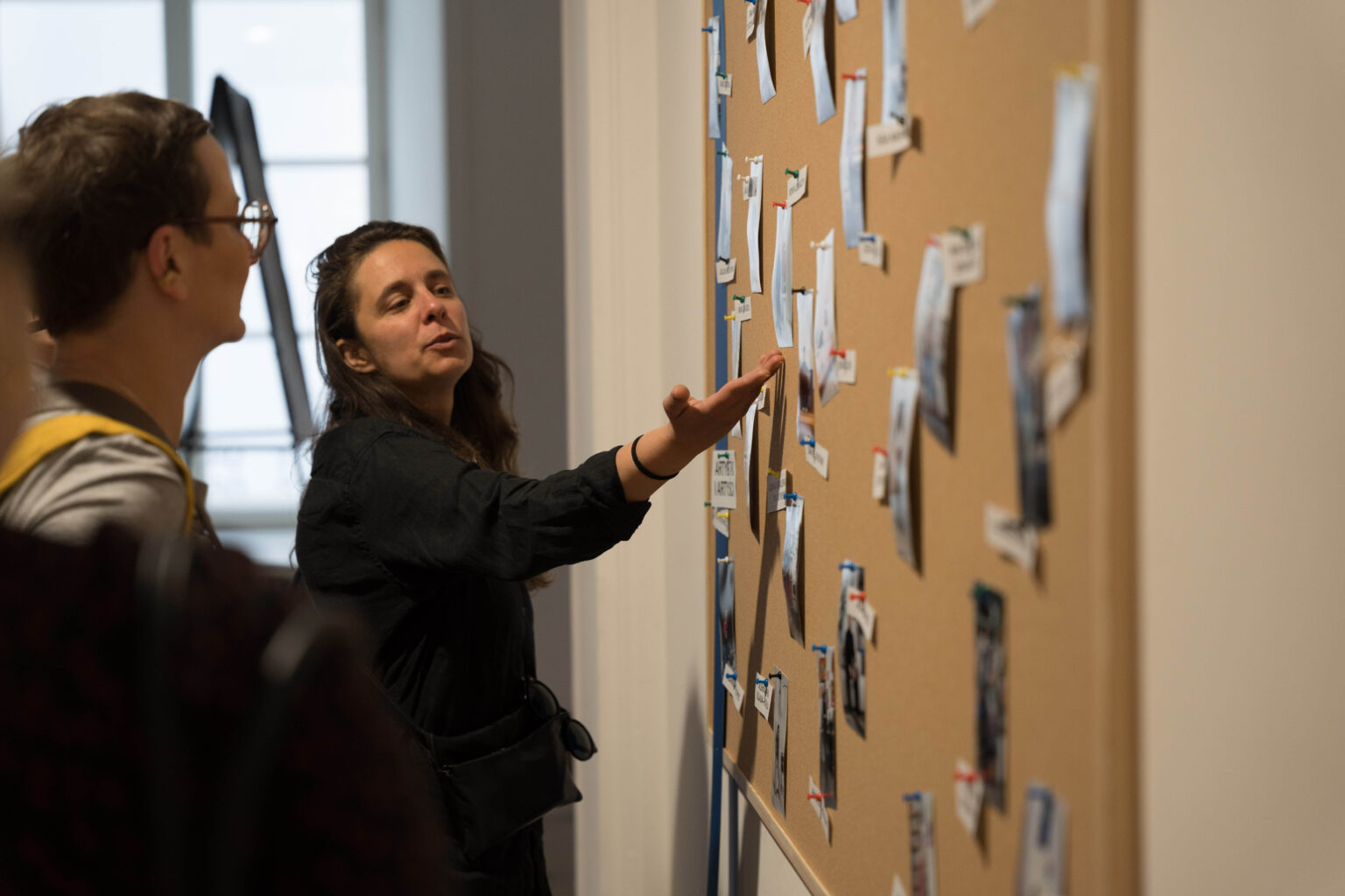 kobieta prezentuje wskazując dłonią nieduże wydruki przyczepione do tablicy korkowej