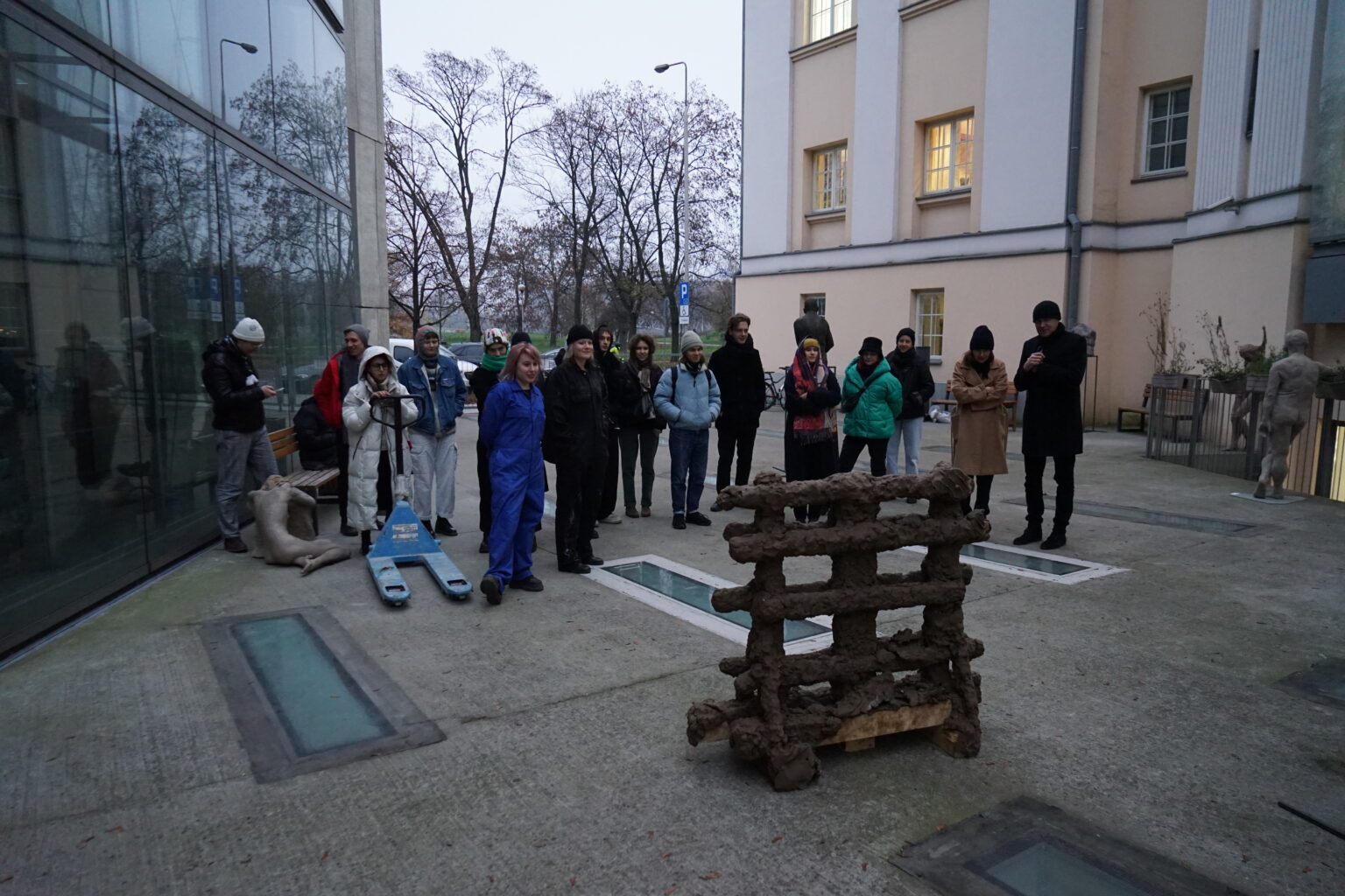 Grupa osób stoi na zewnątrz przed instalacją artystyczną umieszczoną na palecie, pomiędzy dwoma budynkami, w pochmurny dzień.