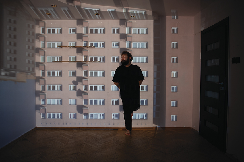 fotografia pzedstawia mężczyznę w czarnym ubraniu opartego plecami o ścianę w pomieszczeniu z widokiem bloków mieszkalnych na ścianie