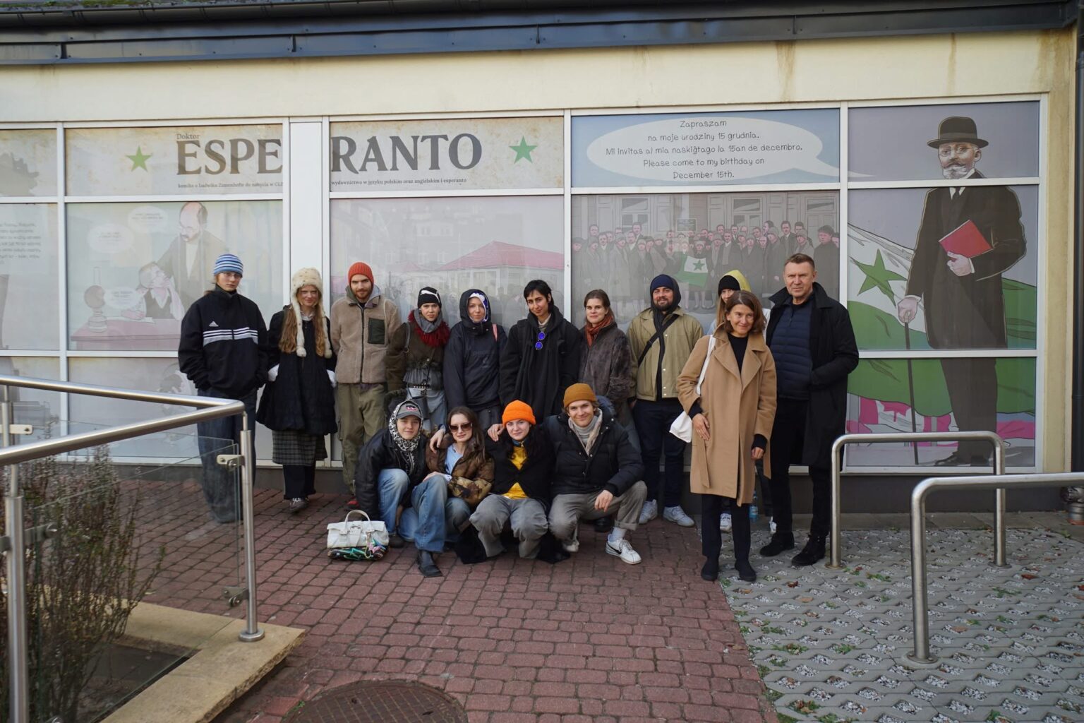 Grupa ludzi pozuje do zdjęcia na zewnątrz budynku z oknami ozdobionymi grafikami i napisem "ESPERANTO".