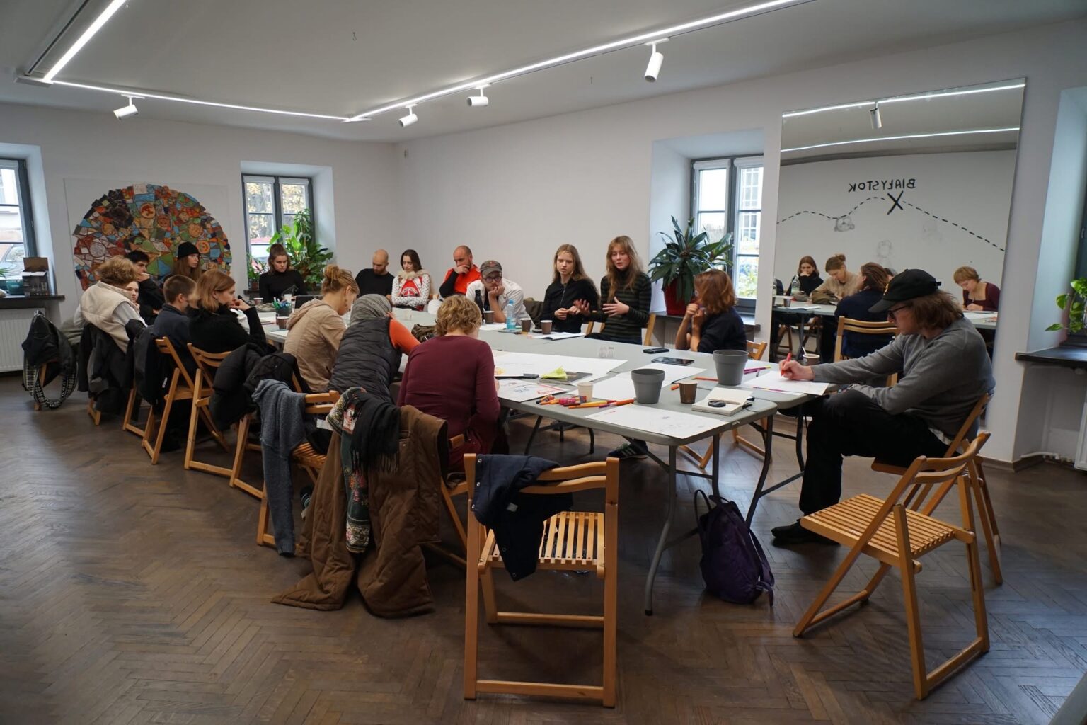 Grupa dorosłych i młodzieży uczestniczących w warsztatach lub spotkaniu przy długim stole w jasnym pomieszczeniu.