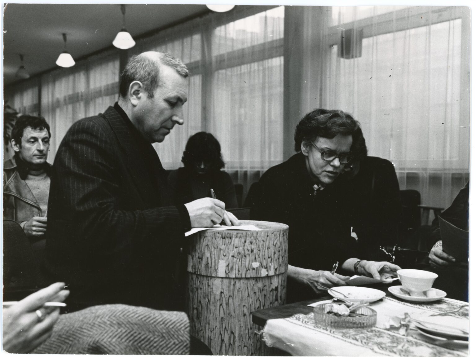 Czarno-białe zdjęcie przedstawia kobietę i mężczyznę w pomieszczeniu o dużych oknach. Wokół ludzie; stół zastawiony filiżankami