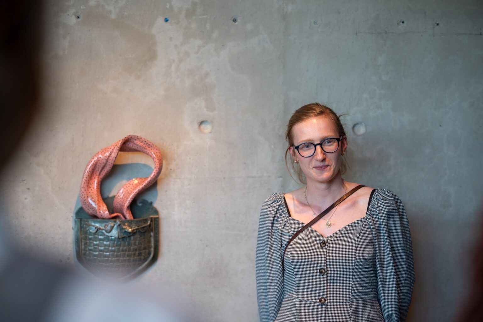 na tle betonowej ściany kobieta w okularach i ceramiczna rzeźba w kształcie damskiej torebki