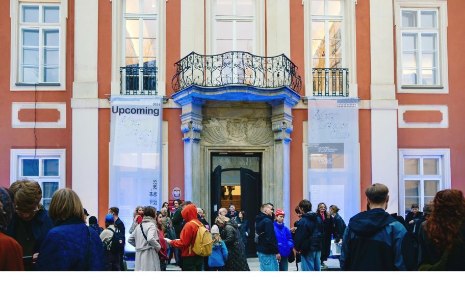 kolorowa fotografia - grupa ludzi przed wejściem do budynku z pomarańczową elewacją