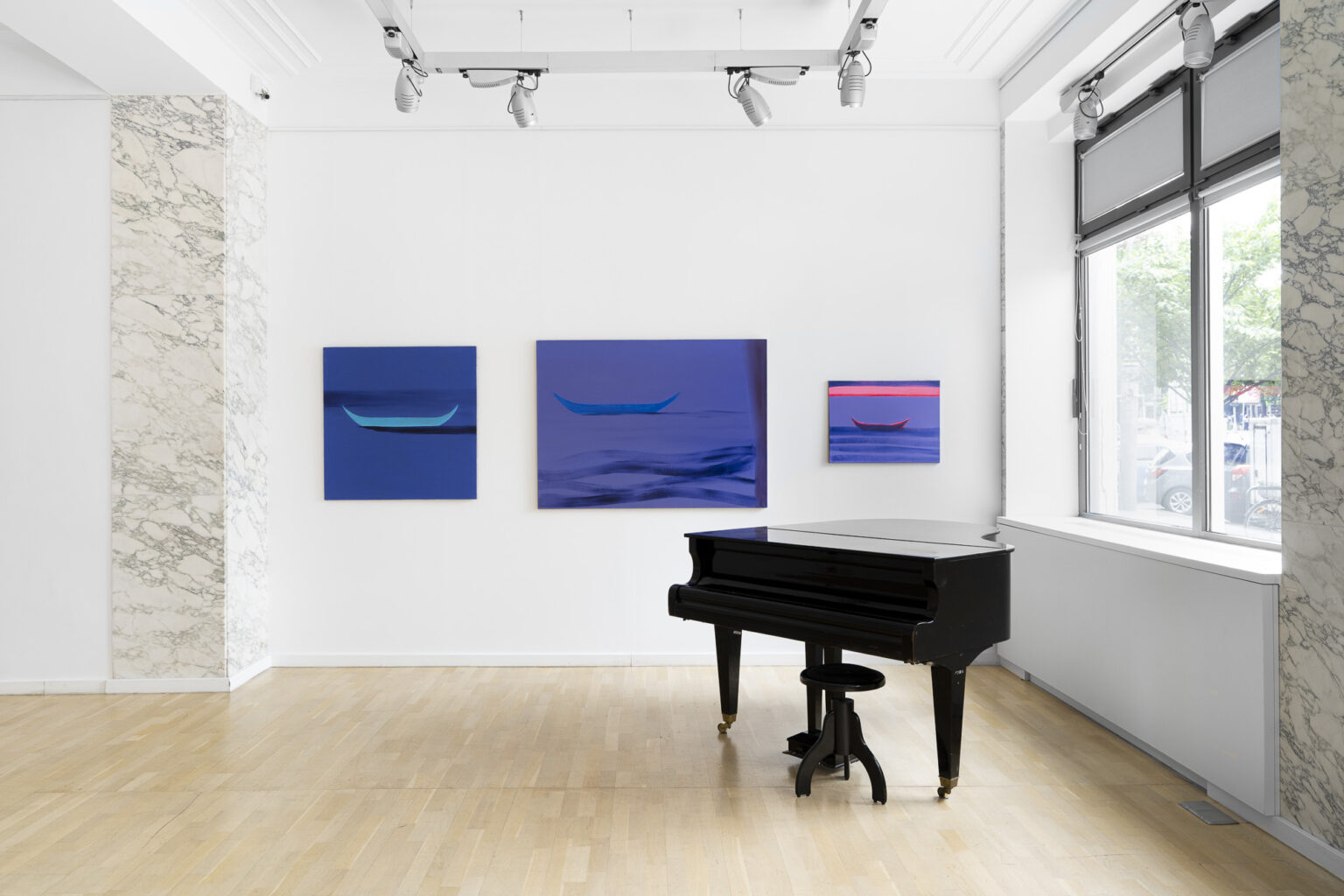 trzy obrazy na ścianie w przestrreni wystawy przedstawiające łódki, po prawe stronie stoi czarny fortepian