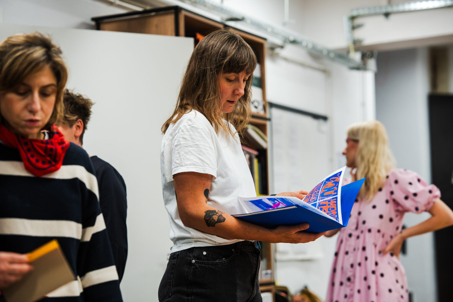 Kobieta w białej koszulce i ciemnych spodniach przegląda niebieski album w pracowni, inne osoby w tle.