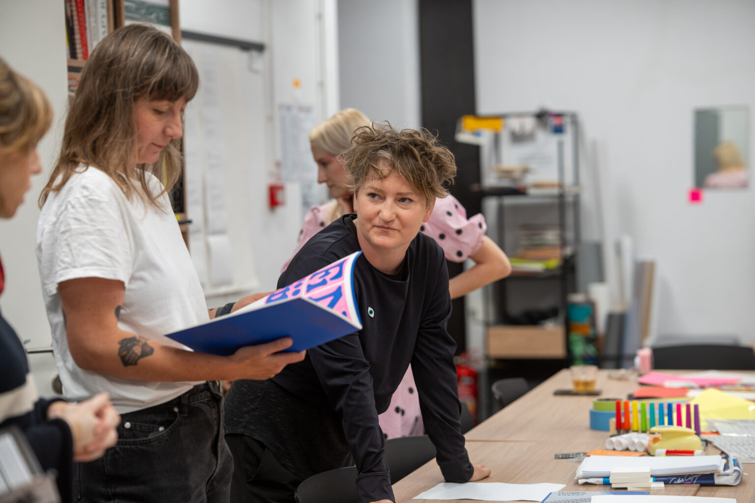 Trzy kobiety w pracowni. Jedna trzyma notes, druga opiera się o stół, trzecia stoi z tyłu. Na stole rozłożone materiały.
