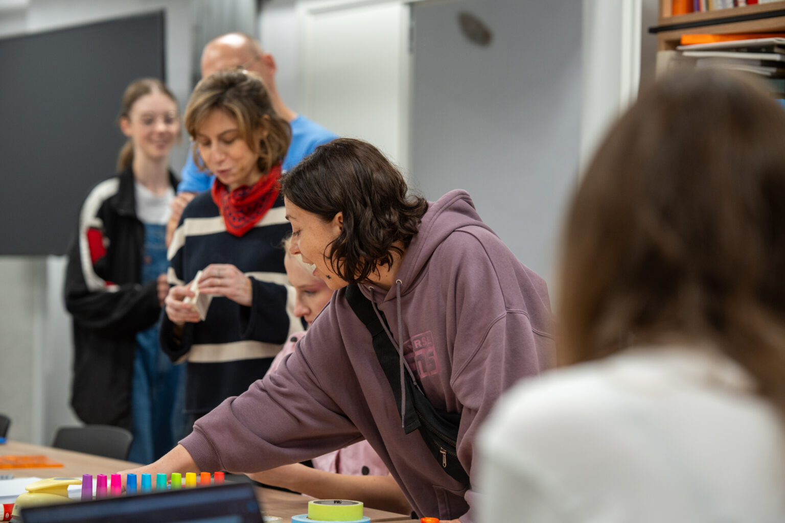 Grupa osób w różnym wieku, pochylających się nad stołem w pracowni, angażuje się w twórcze działanie, skupiając się na detalach.