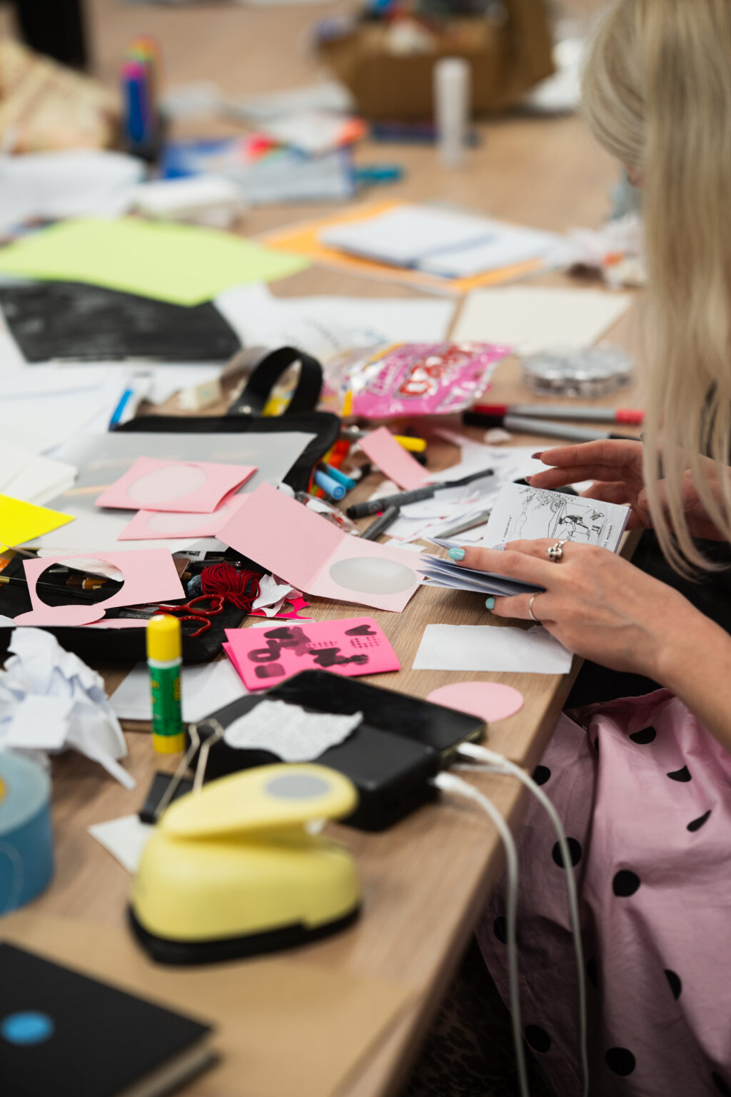 Blondwłosa osoba pracuje przy stole pełnym artykułów papierniczych: nożyczek, kleju, kolorowego papieru i dziurkacza.