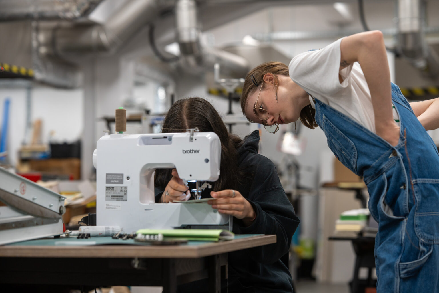 Dwie osoby w pracowni, jedna pochylona nad maszyną do szycia, a druga patrzy na nią, w tle widać wnętrze warsztatu.