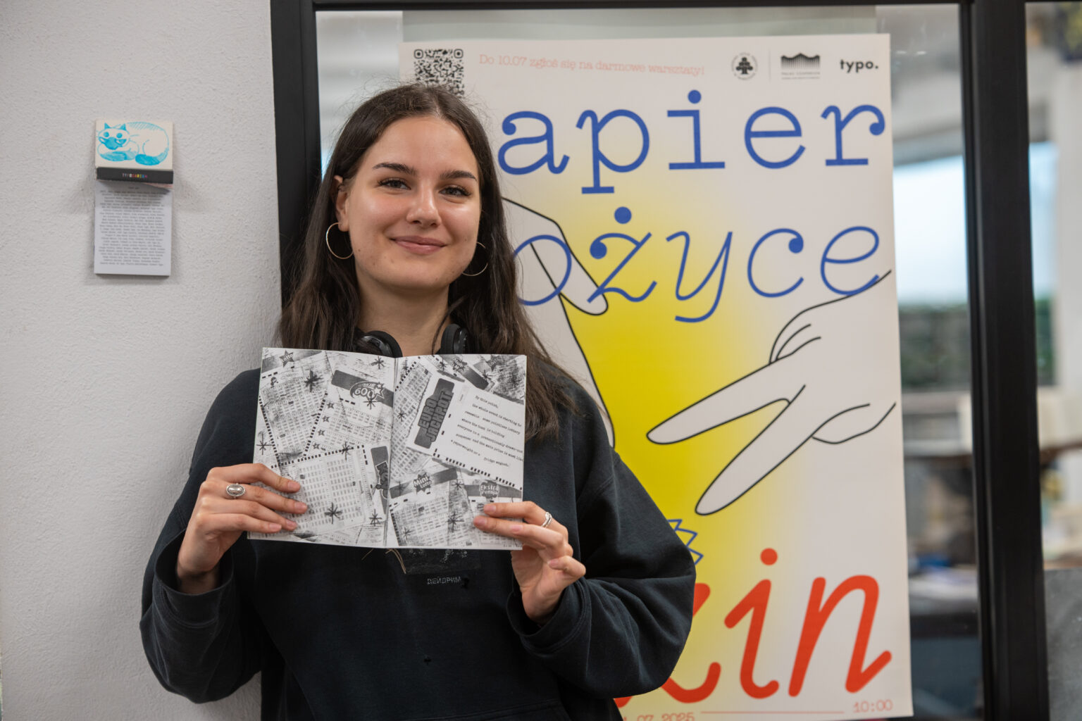 Młoda kobieta uśmiecha się, trzymając otwarty zin przed plakatem "papier nożyce zin" z grafiką dłoni.
