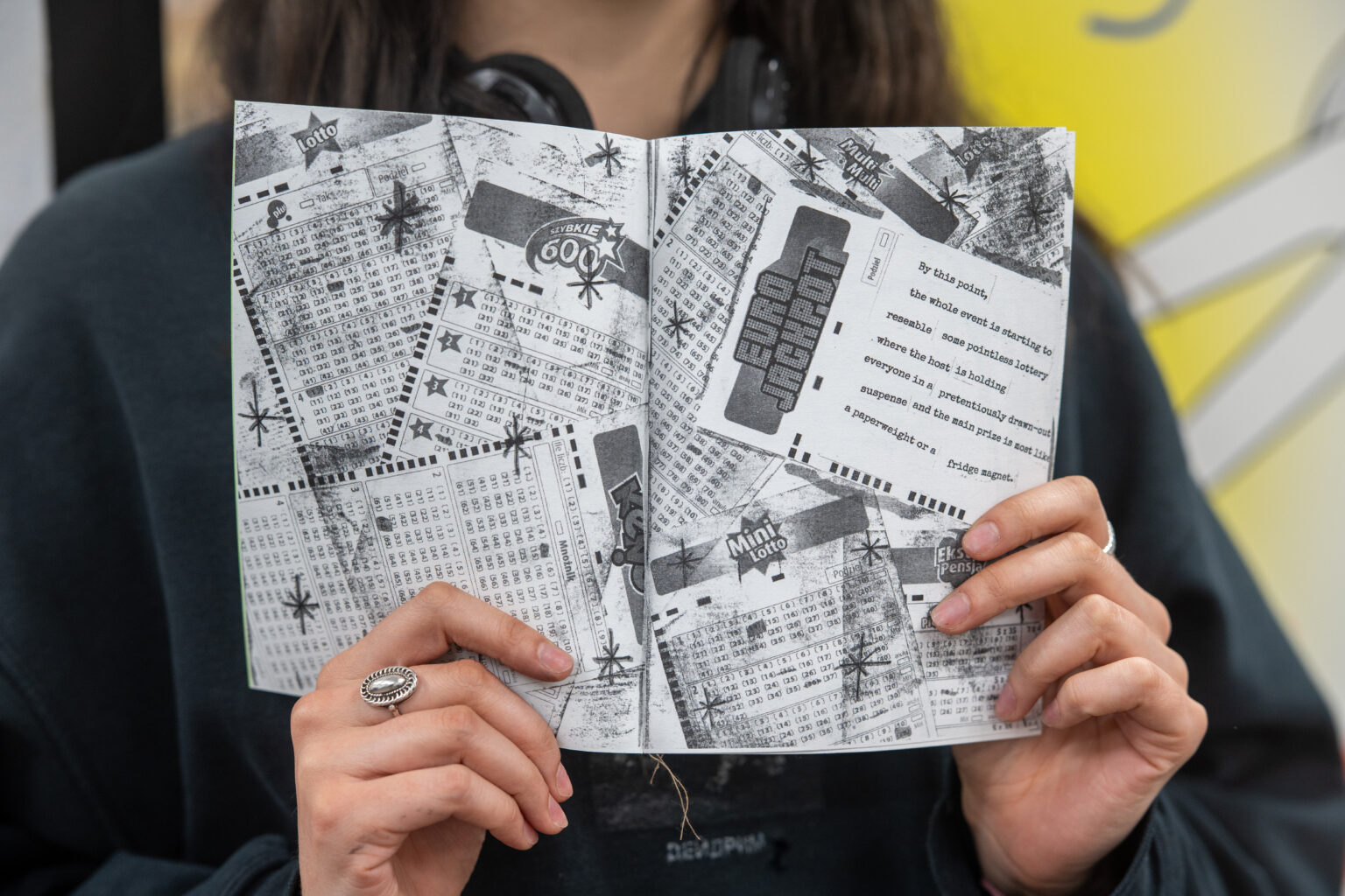 Osoba trzyma otwarty, czarno-biały zin, którego strony pokrywają kolaże z tekstem, liczbami i grafikami o tematyce biurokratycznej.