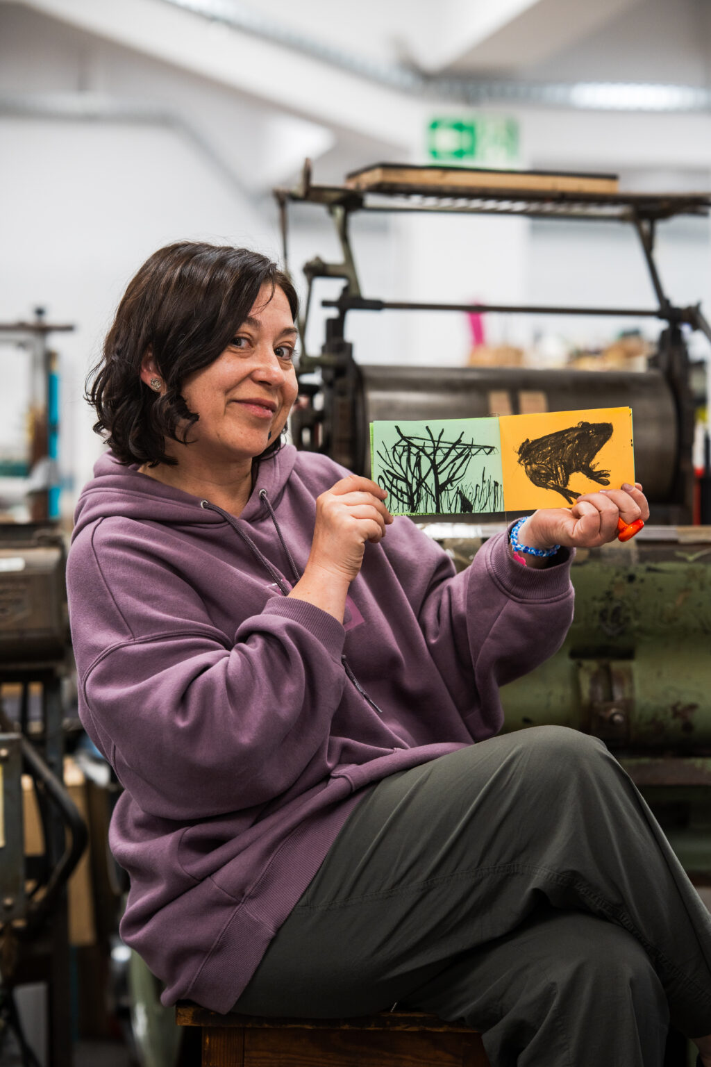 Kobieta siedząca na krześle uśmiecha się, prezentując dwie odbitki, jedna z drzewami, druga z żabą, w tle maszyny drukarskie.