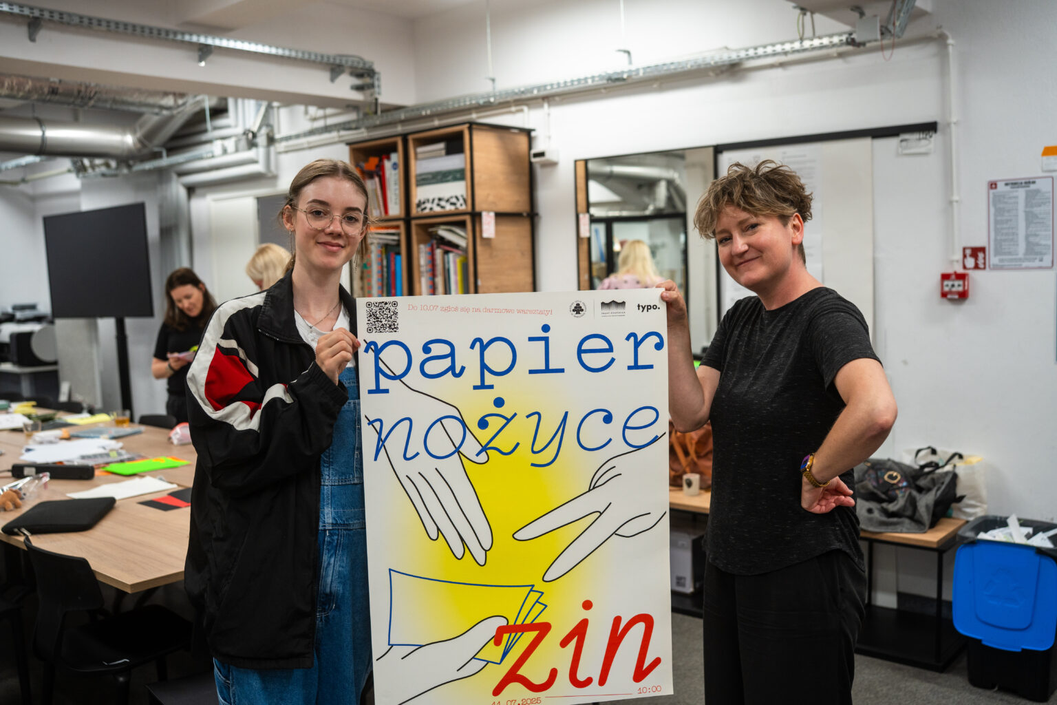 Dwie kobiety trzymają plakat z napisem "papier, nożyce, zin" i ilustracjami dłoni, w pracowni z regałami i stołami.