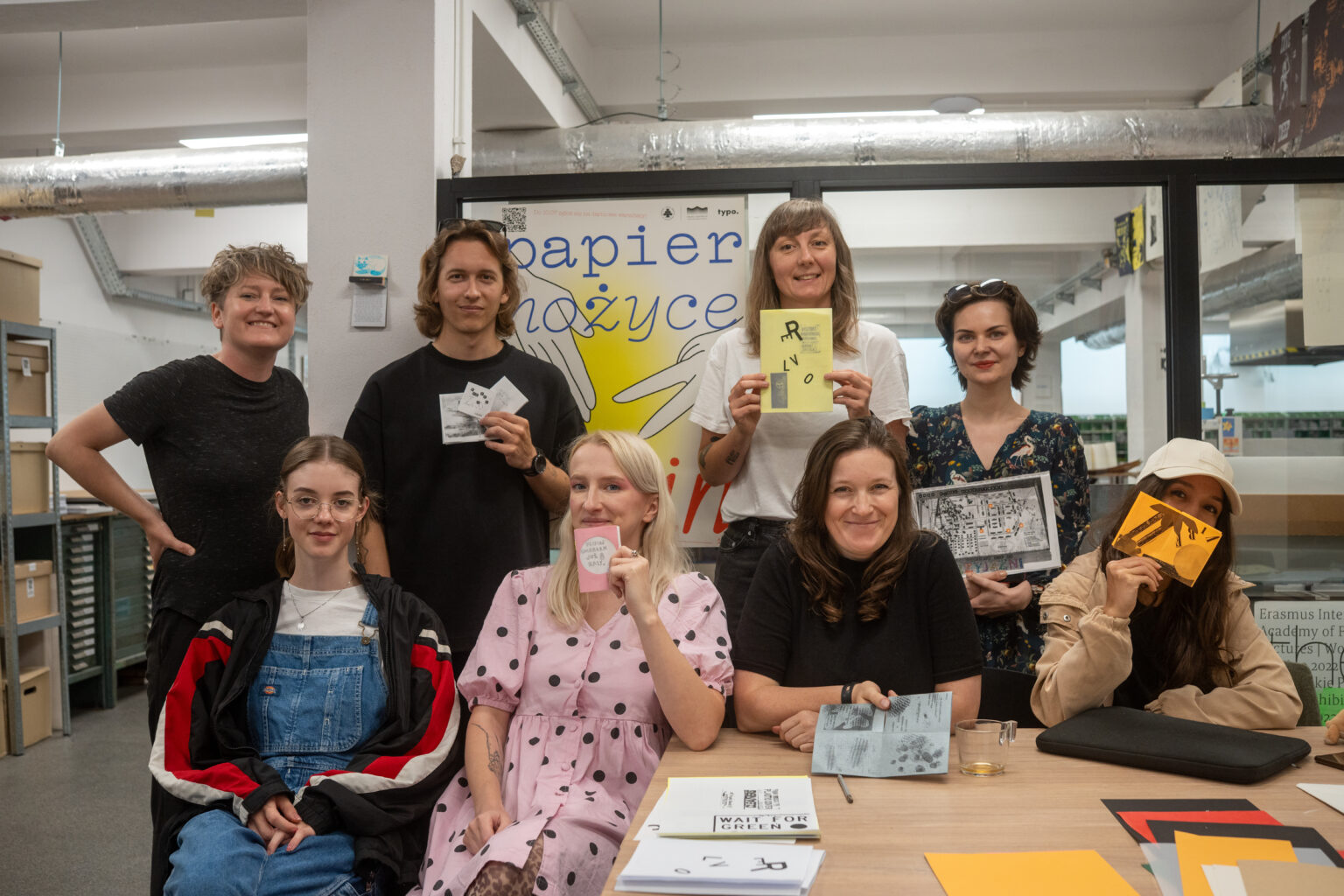Grupa ośmiu osób w różnym wieku, stojących i siedzących, prezentuje swoje prace w pracowni, w tle plakat "papier nożyce".