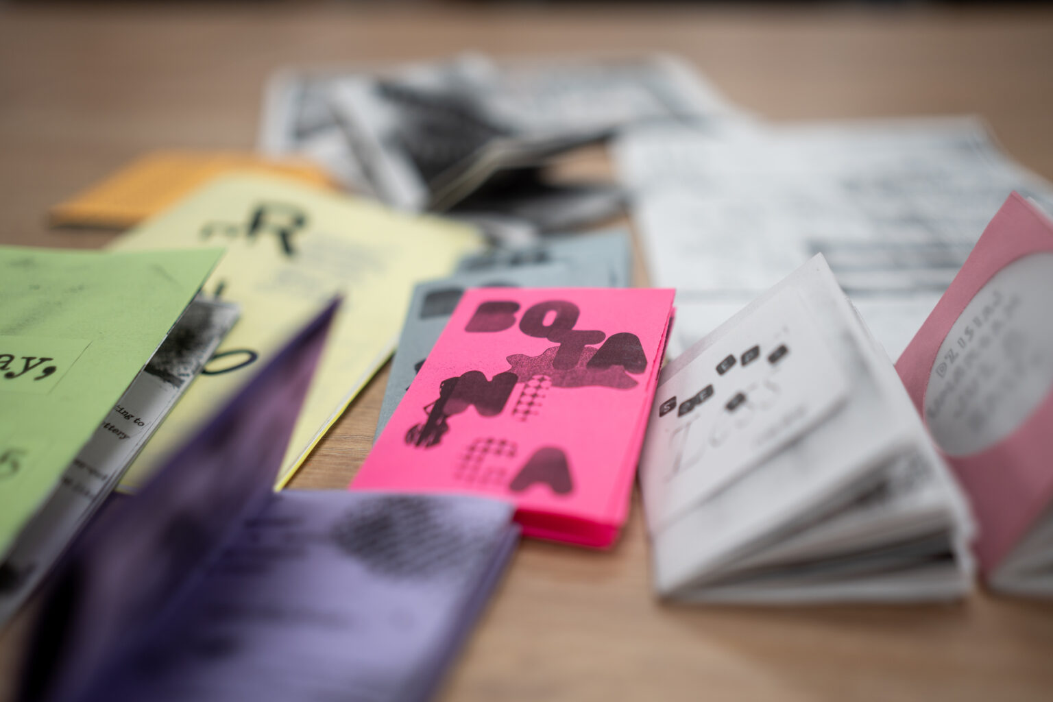 Zbliżenie na różnorodne, kolorowe, ręcznie wykonane broszury i ziny leżące na drewnianym stole, niektóre otwarte, z tekstem i grafikami.