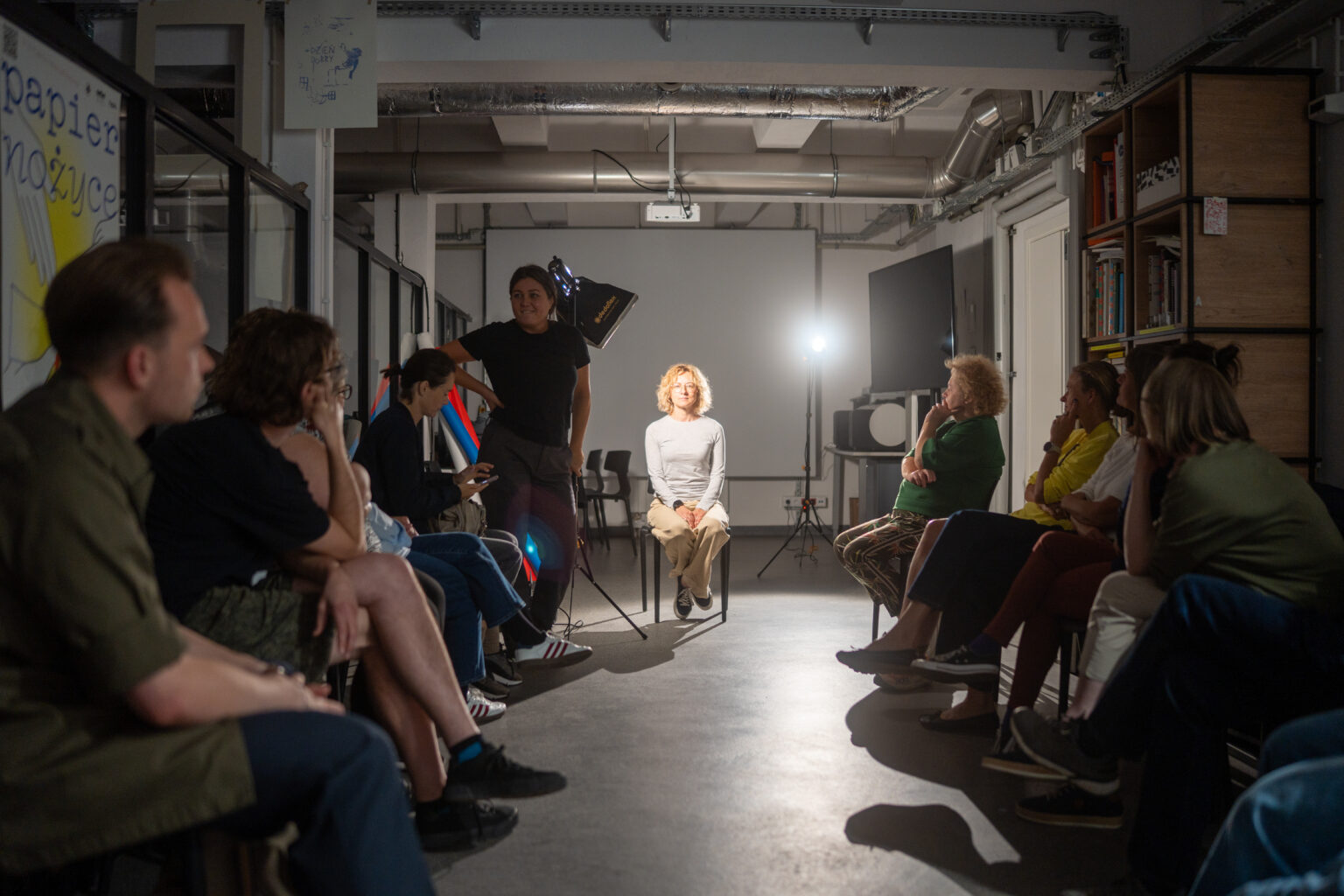 Ludzie siedzą w półkolu i oglądają kobietę siedzącą na krześle, oświetloną lampą.