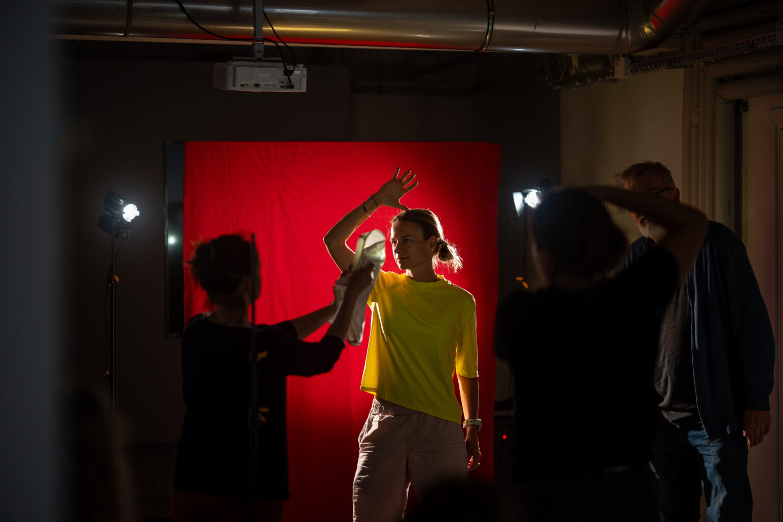 Kobieta pozuje przed czerwonym tłem. Ktoś inny pomaga jej, trzymając coś w ręku.