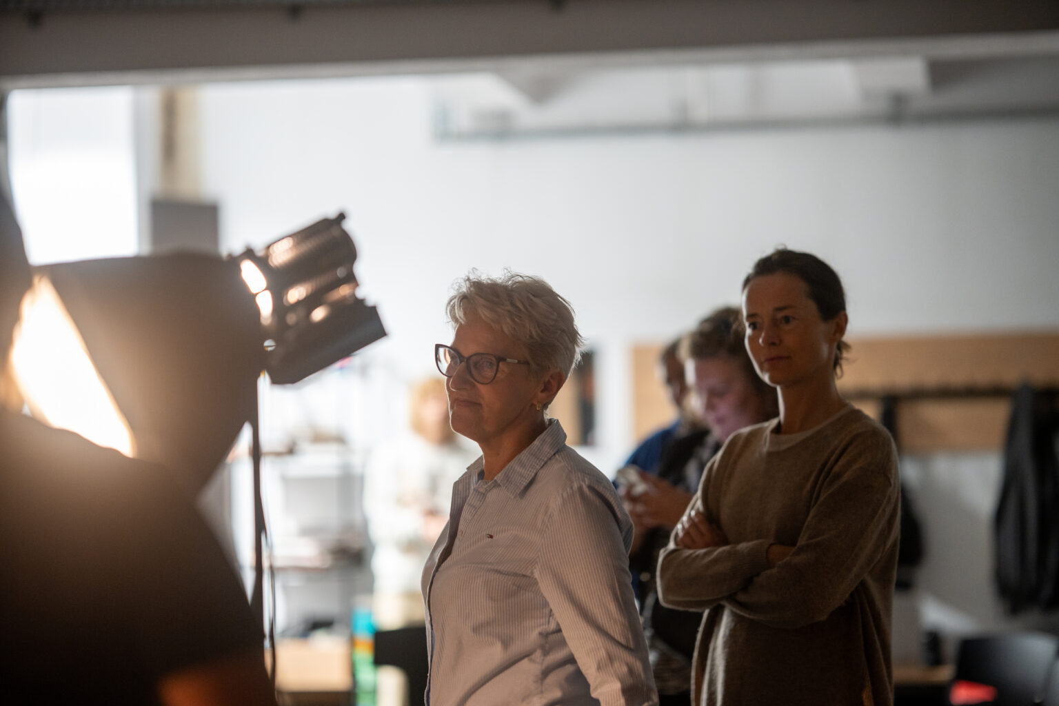 Dwie kobiety stoją obok siebie i patrzą w prawo. W tle widoczne są lampy studyjne.