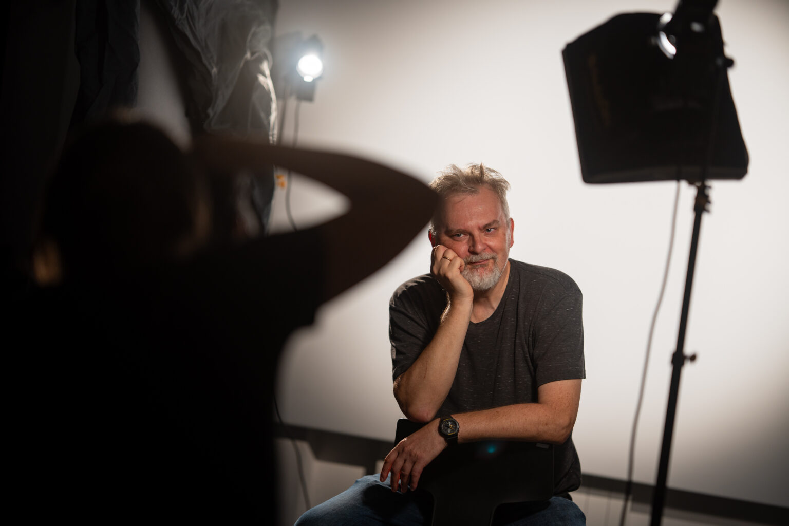 Mężczyzna uśmiecha się, siedząc na krześle, podczas sesji zdjęciowej. W tle widoczne jest oświetlenie.