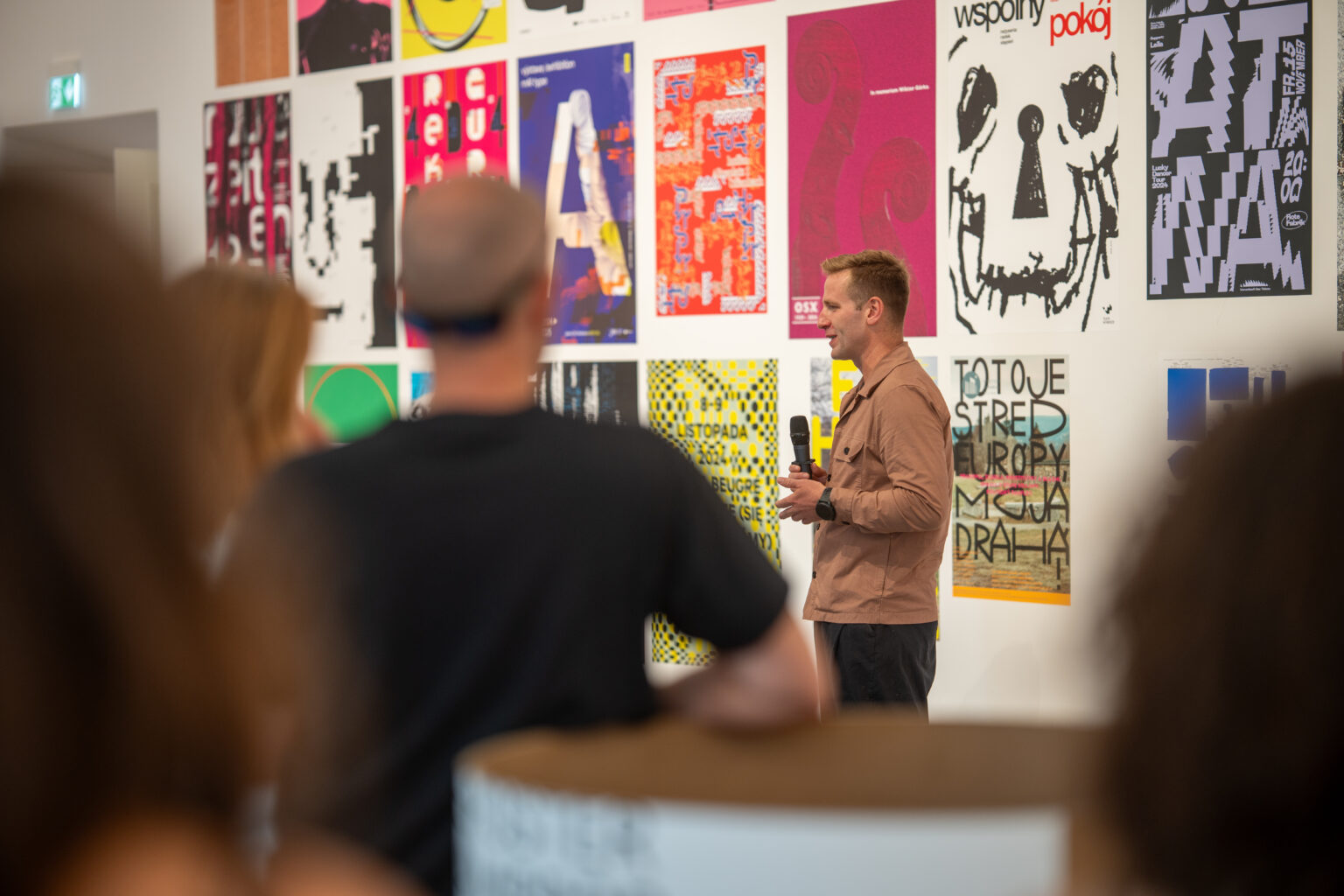 Mężczyzna rozmawia z ludźmi w galerii sztuki, na ścianach za nim wiszą plakaty.