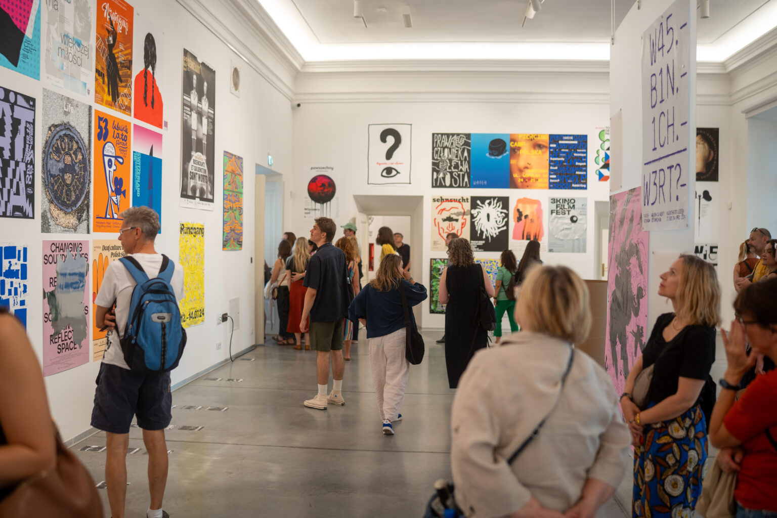 Ludzie oglądają plakaty i obrazy w galerii sztuki. Na ścianach widać wiele kolorowych dzieł.