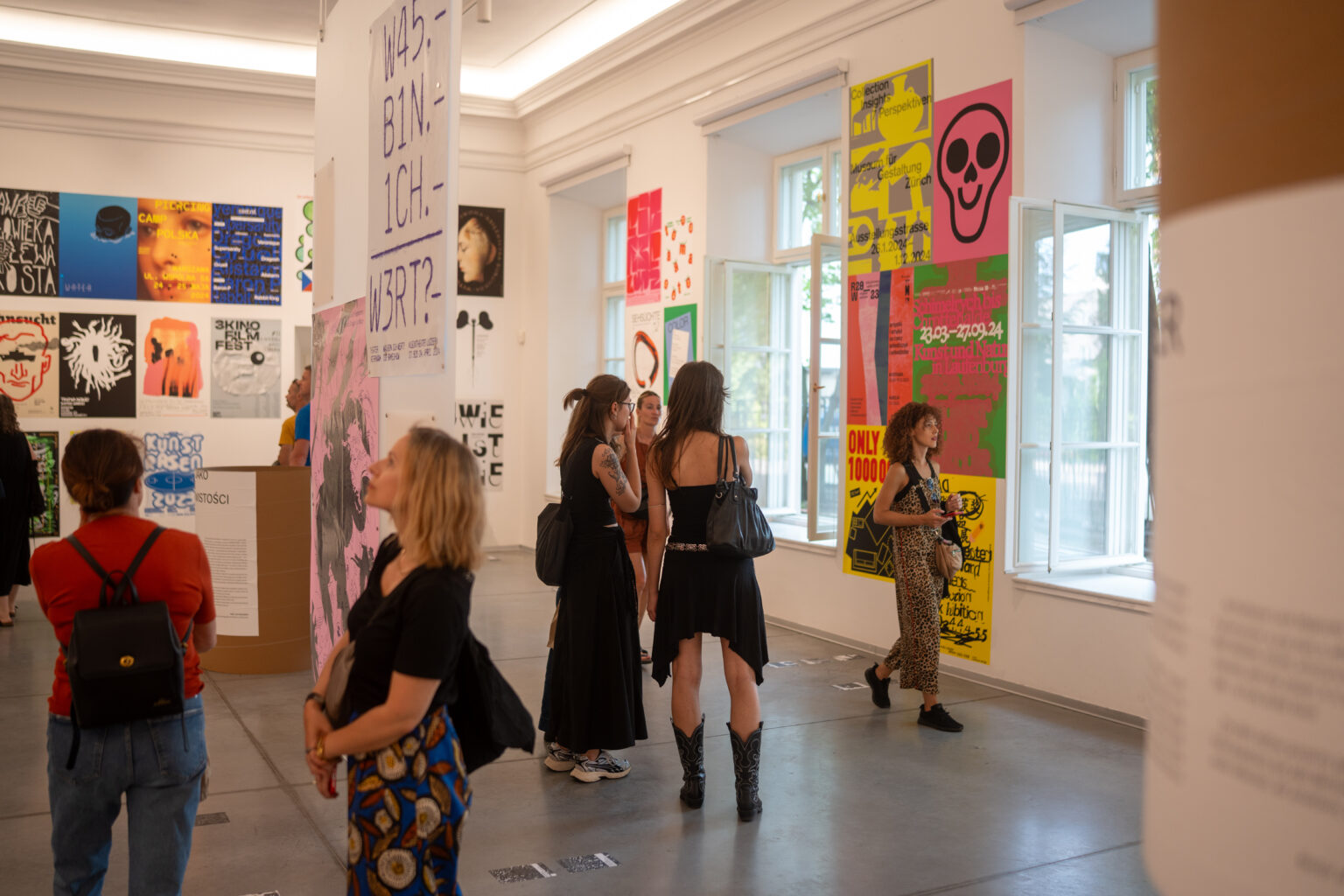 Ludzie oglądają plakaty i grafiki na ścianach w jasnej, przestronnej galerii.