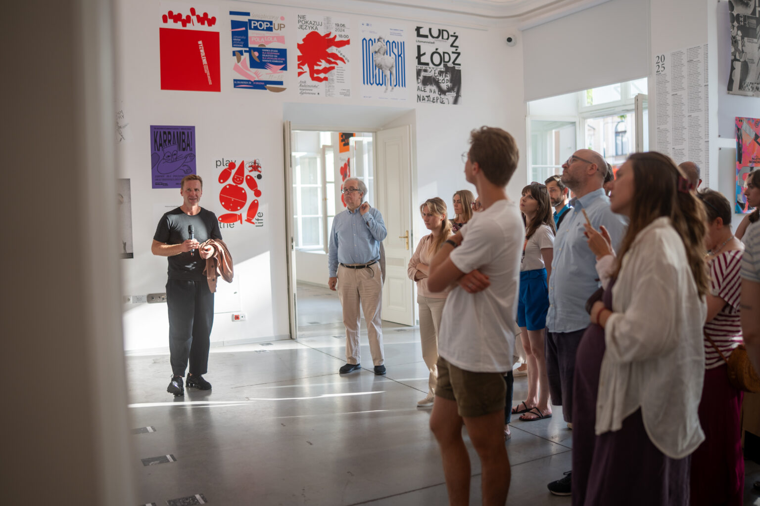 Ludzie oglądają wystawę sztuki. Na ścianach widać plakaty w różnych stylach i kolorach.