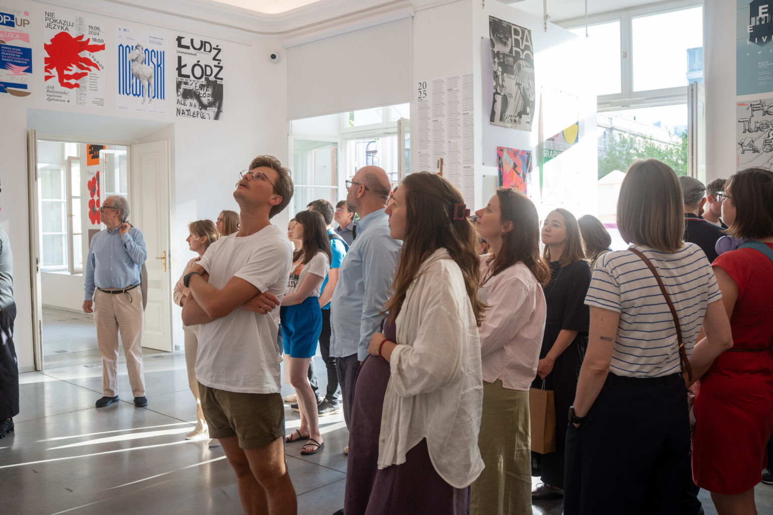 Grupa ludzi patrzy w górę podczas wystawy sztuki. Na ścianach i suficie wiszą plakaty.