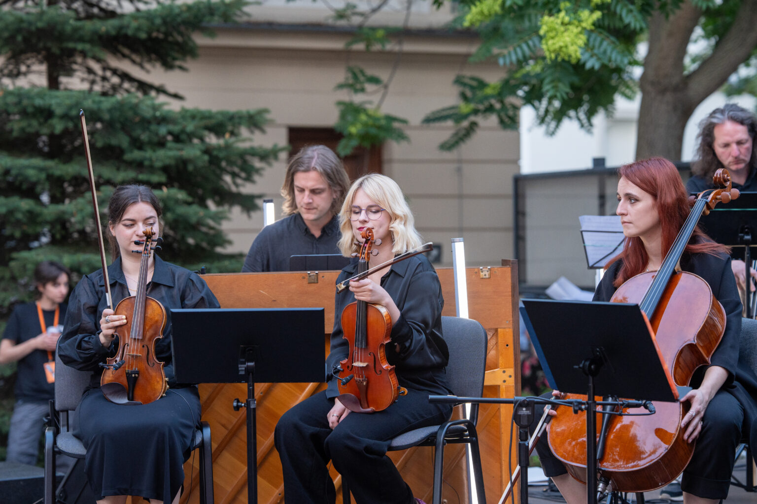 Muzycy z orkiestry smyczkowej grają na świeżym powietrzu. Dwie kobiety grają na skrzypcach, a trzecia na wiolonczeli.