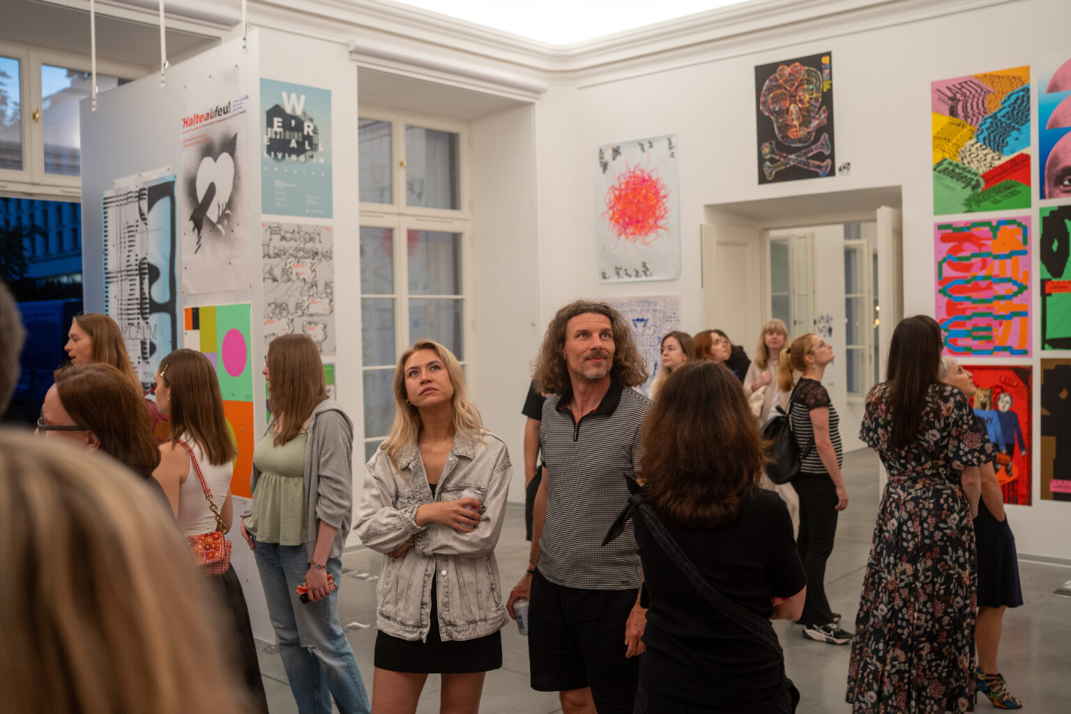 Ludzie oglądają dzieła sztuki. W tle widać plakaty i obrazy w różnych stylach i kolorach.