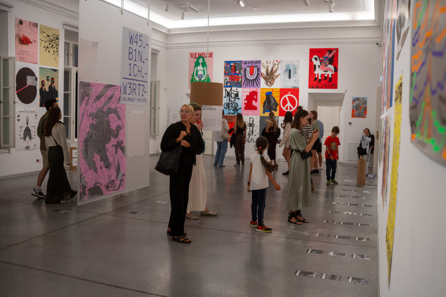 Ludzie oglądają dzieła sztuki w galerii. Na ścianach wiszą kolorowe obrazy.