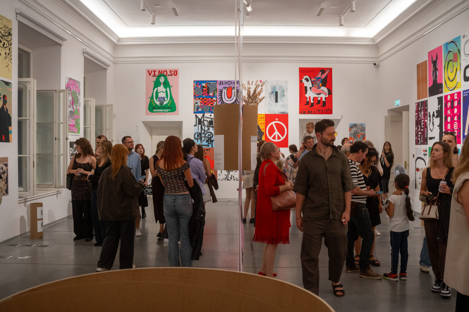 Ludzie w jasnym pomieszczeniu oglądają plakaty i grafiki na ścianach galerii. Widać prace z symbolem pokoju i innymi motywami.