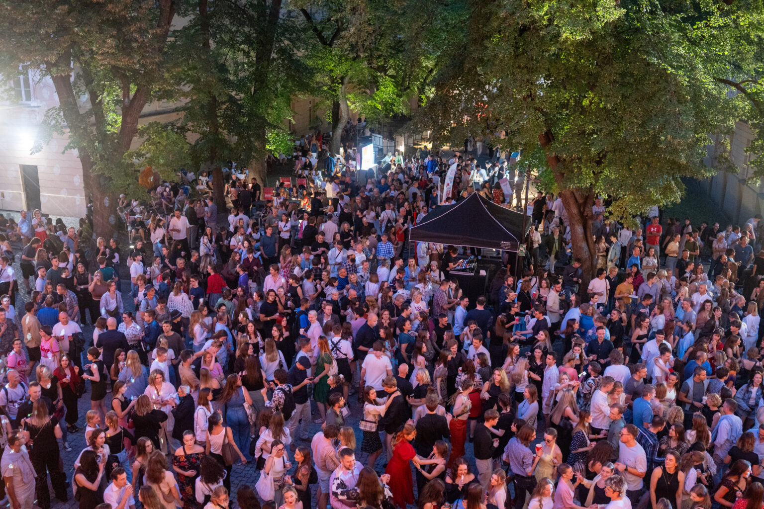 Ogromny tłum ludzi na koncercie na świeżym powietrzu. Na środku placu widoczna jest scena lub stoisko z czarnym dachem.