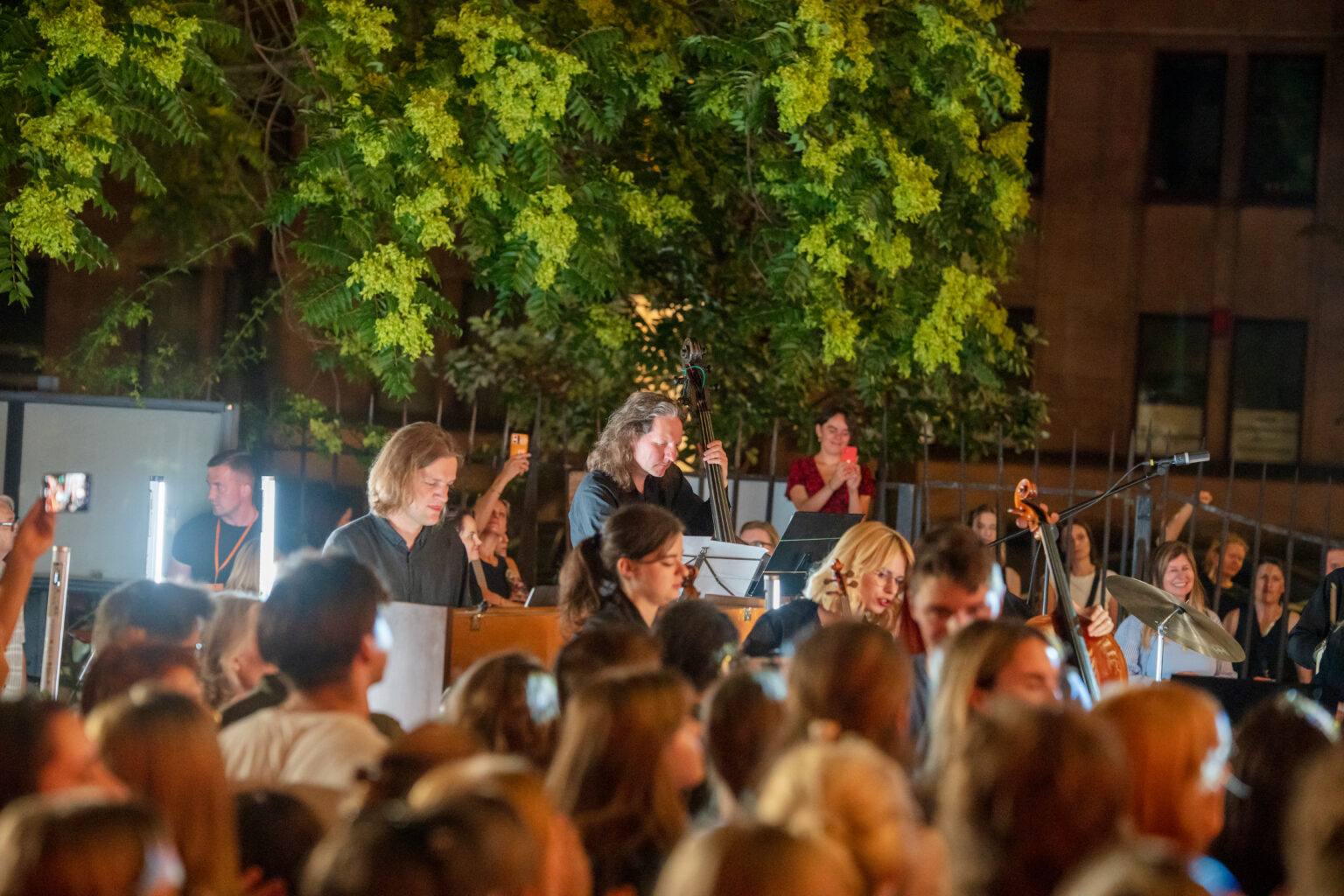 Orkiestra na scenie podczas koncertu na świeżym powietrzu. Ludzie w tłumie oglądają występ, rozmawiają i robią zdjęcia.