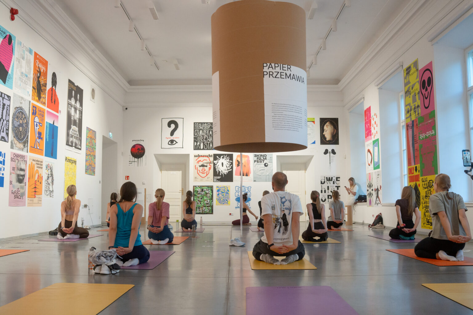 Ludzie siedzą na matach na podłodze, ćwicząc jogę w galerii sztuki. Na ścianach widać plakaty.