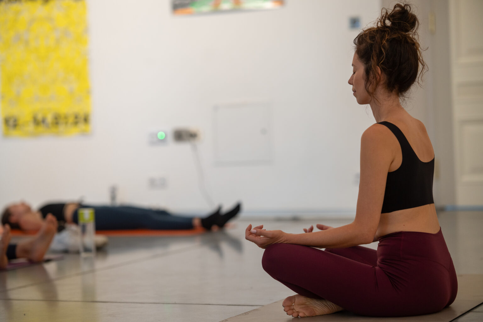 Kobieta siedzi w pozie medytacyjnej na macie do jogi. Inni ludzie leżą w tle.