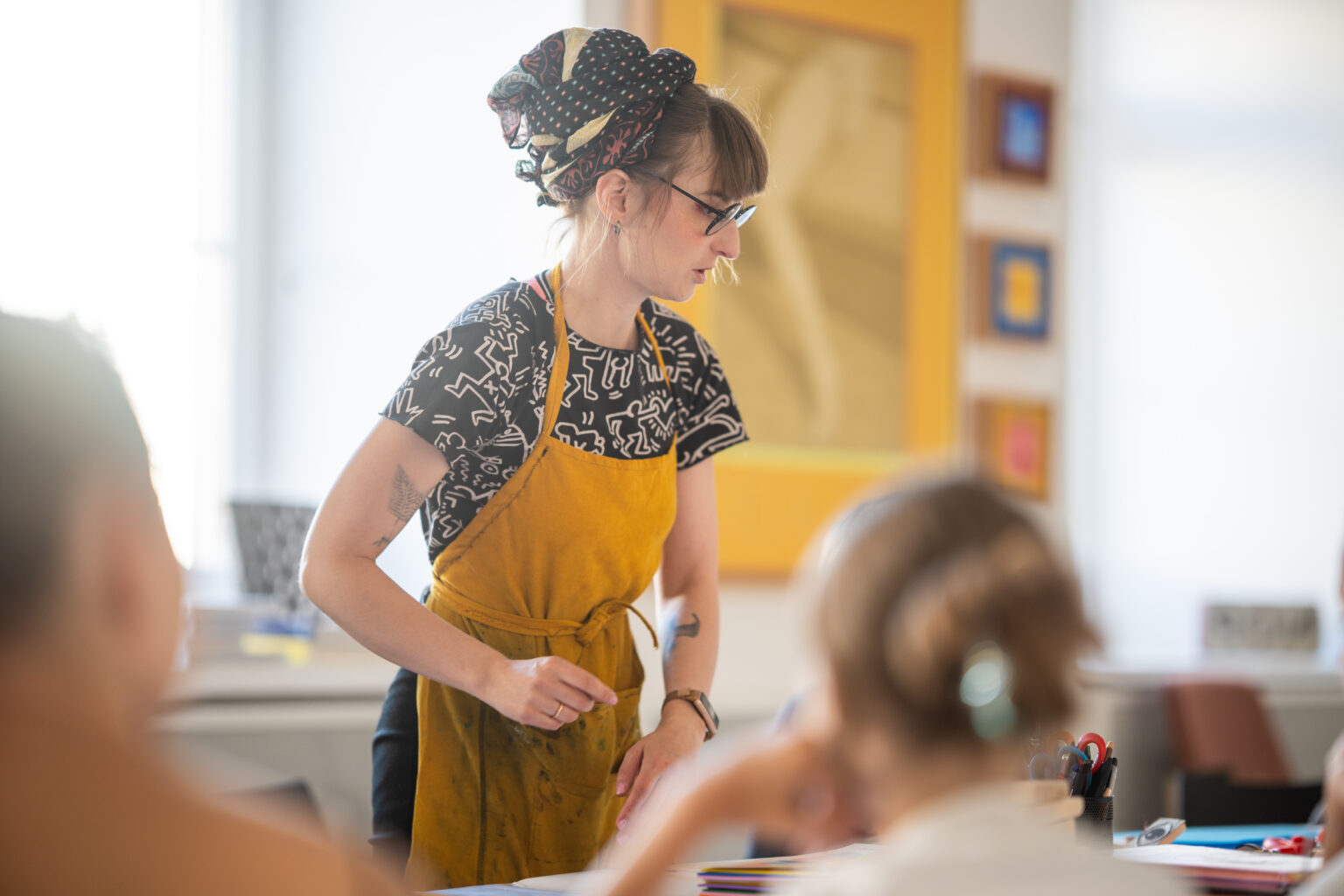 Kobieta w okularach i żółtym fartuchu pomaga dzieciom podczas warsztatów artystycznych.