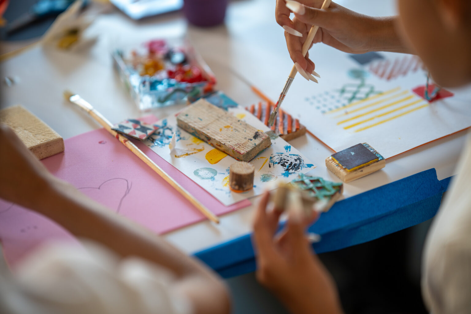 Dzieci malują i tworzą odbitki z pomocą pędzli i gąbek. W tle widać farby i przybory plastyczne.