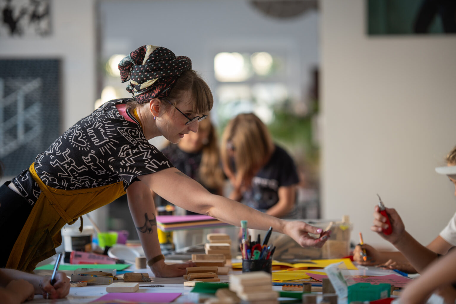 Kobieta w okularach i chustce na głowie pomaga dziecku podczas warsztatów artystycznych. Na stole leżą materiały plastyczne.
