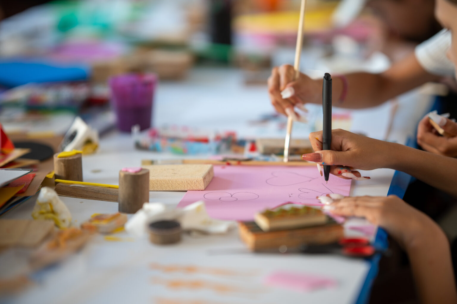 Dzieci siedzą przy stole i uczestniczą w warsztatach plastycznych. Rysują i malują na różowym papierze.