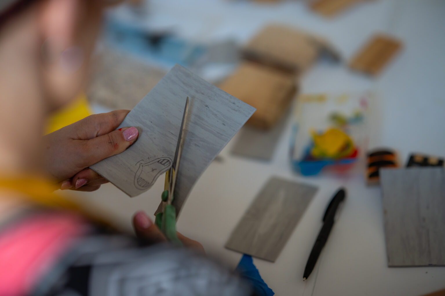 Ktoś trzyma nożyczki i wycina szary papier. Na stole widać inne materiały do warsztatów plastycznych.