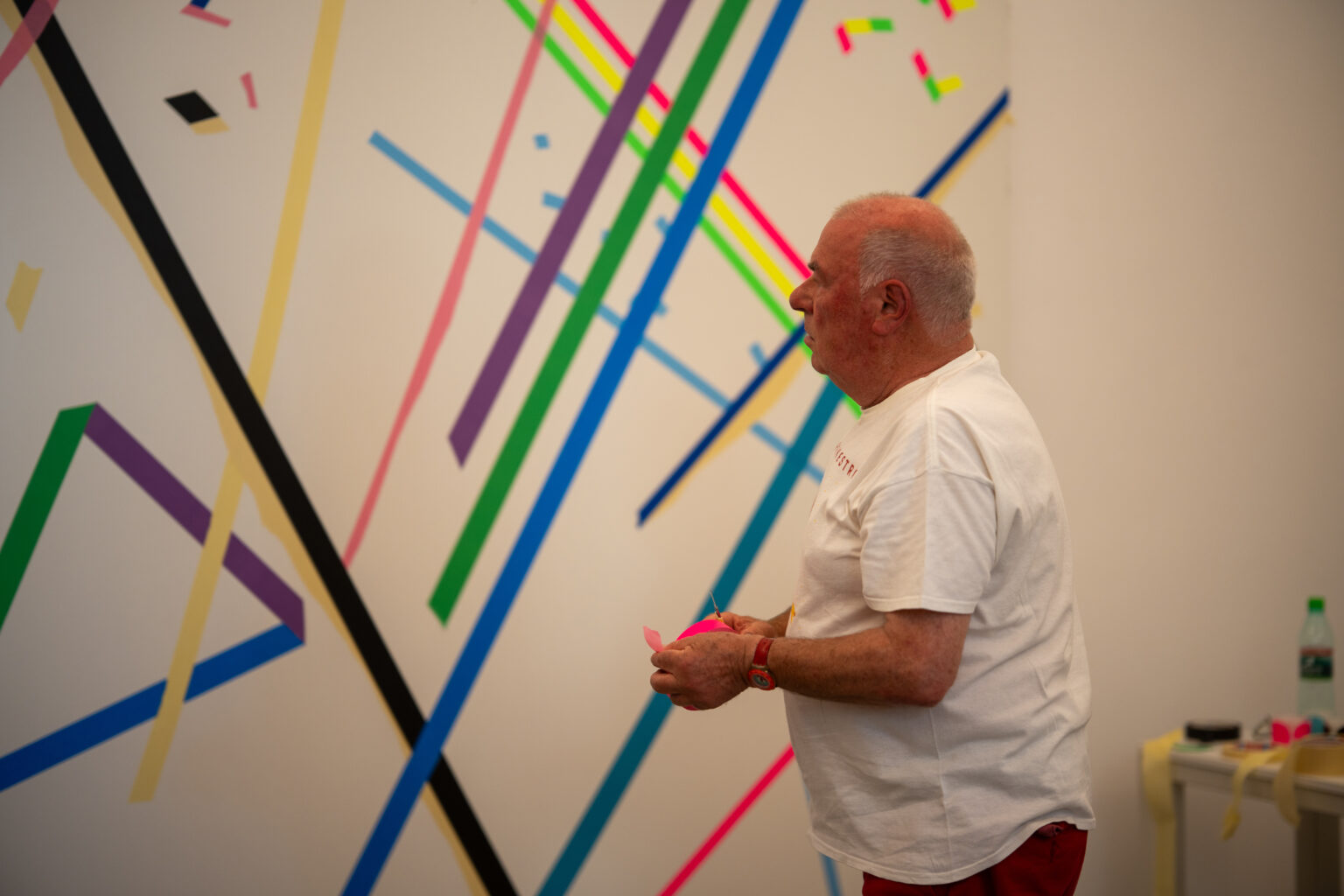Mężczyzna z siwą głową, ubrany w białą koszulkę i czerwone spodnie, stoi przed abstrakcyjnym obrazem z kolorowych taśm