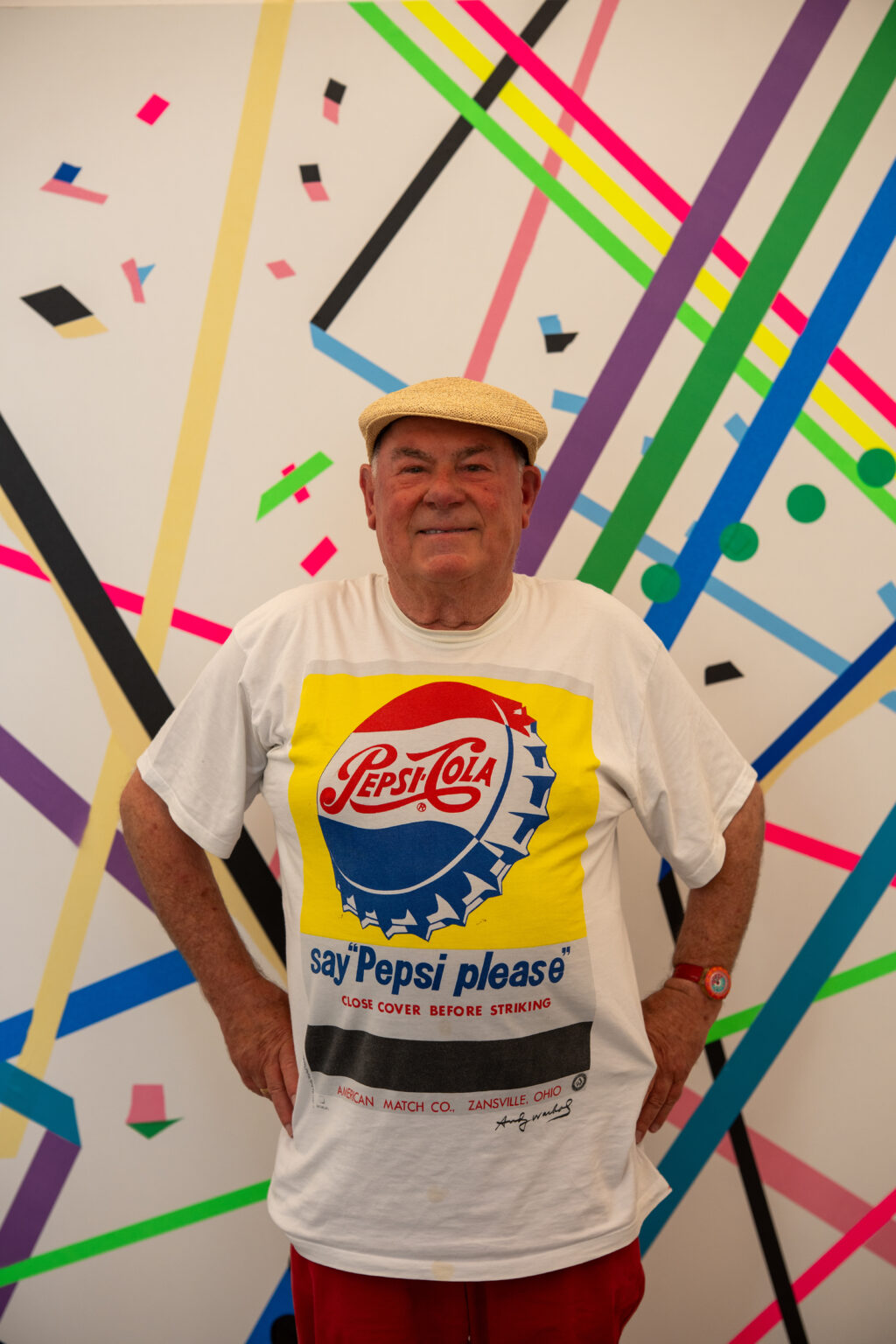 Mężczyzna w jasnym kaszkiecie i białej koszulce z logo Pepsi-Cola stoi przed abstrakcyjną ścianą z kolorowymi geometrycznymi kształtami.