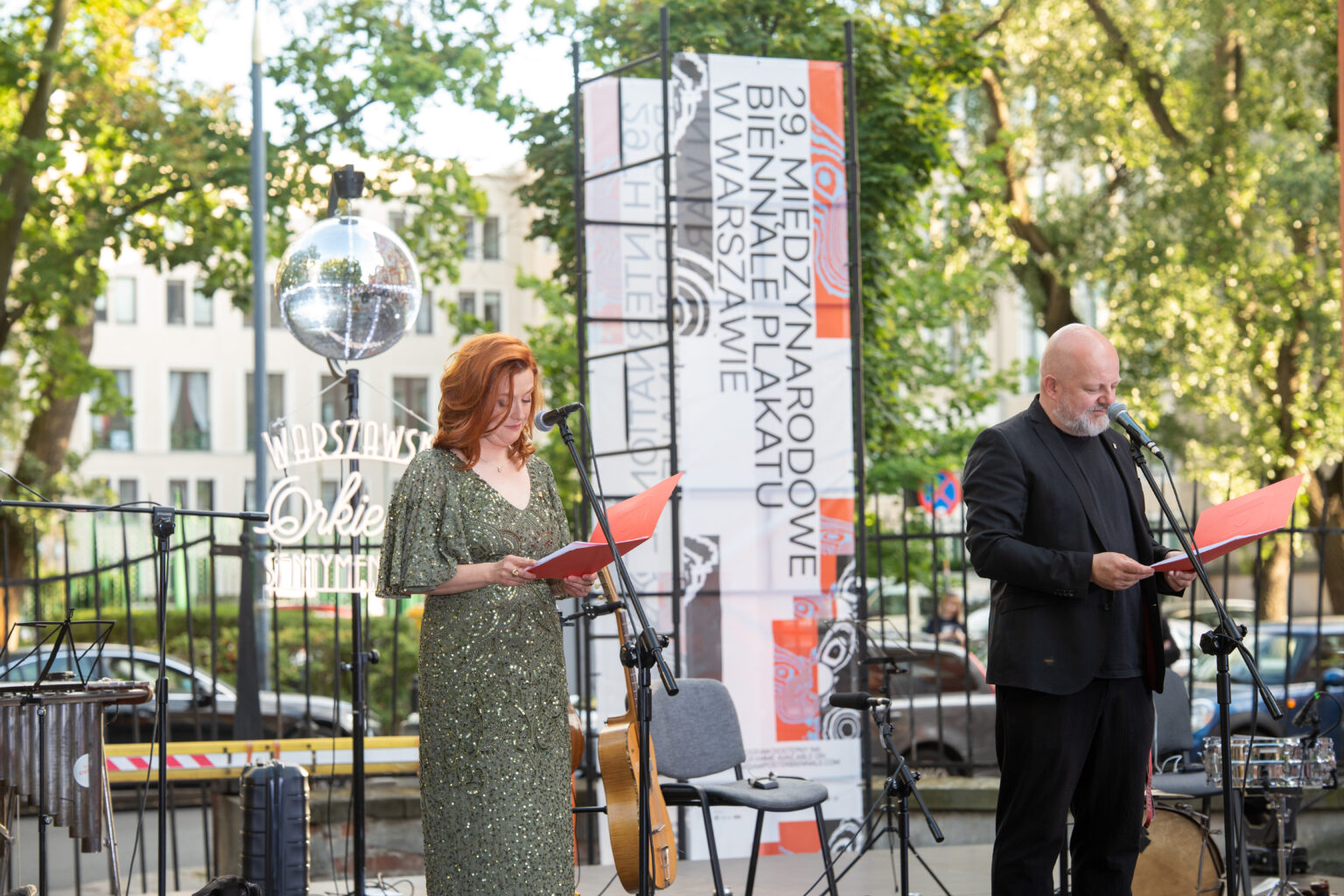 Kobieta w zielonej sukni i mężczyzna w czarnej koszuli czytają z czerwonych teczek na scenie, obok baneru "Międzynarodowe Biennale Plakatu".
