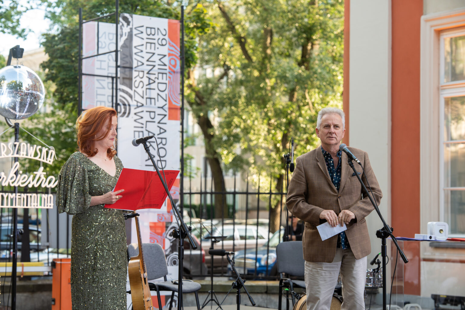 Uśmiechnięta kobieta w cekinowej sukni przemawia do publiczności obok uśmiechniętego mężczyzny w marynarce.