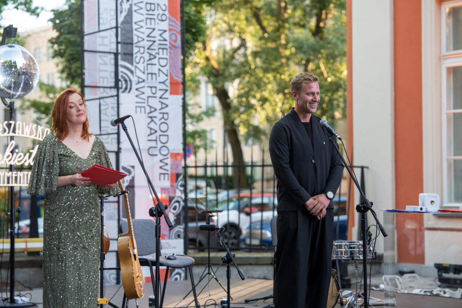 Kobieta w zielonej sukni czyta z czerwonej teczki, a uśmiechnięty mężczyzna w czarnej koszuli stoi obok niej na scenie.